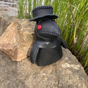 May include: Black leather plague doctor doll with a black hat and red button eyes. The doll is sitting on a rock.