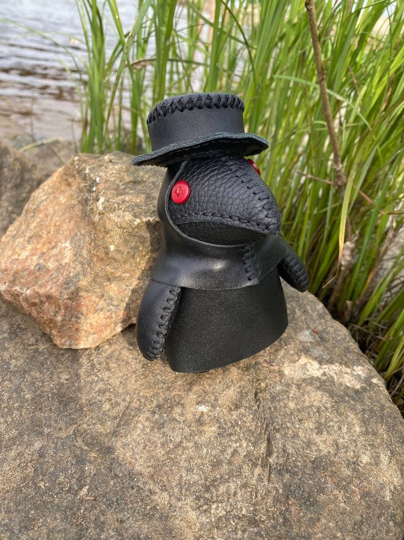 May include: Black leather plague doctor doll with a black hat and red button eyes. The doll is sitting on a rock.