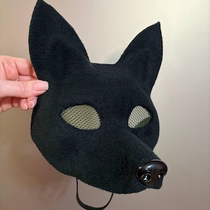 May include: A black dog mask with large, upright ears and a rounded snout. The eye openings are covered with a tan mesh material. A black strap is attached to the mask. The mask is held by a hand.