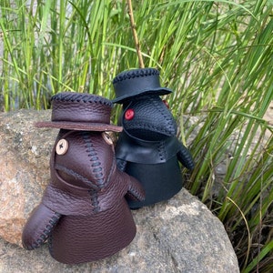 May include: Two small, handmade leather dolls dressed as plague doctors. The doll on the left is brown with a brown hat and the doll on the right is black with a black hat. Both dolls have red button eyes.