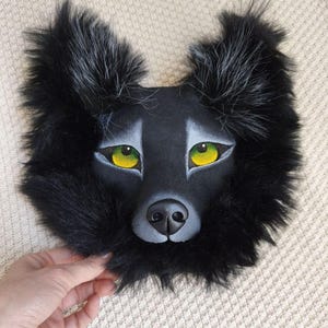 Pu&ograve; includere: Una maschera da lupo nera con pelo lungo e scuro lungo il bordo, orecchie a punta con peli argentati e occhi giallo-verdi brillanti. Il viso presenta una finitura nera opaca e liscia, arricchita da dettagli grigi intorno agli occhi e al naso.