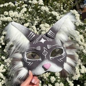 May include: A grey and white cat mask with a pink nose and gold mesh eye coverings. The mask has white accents and fluffy white and grey fur around the ears and cheeks. The mask is held in front of a background of white flowers.