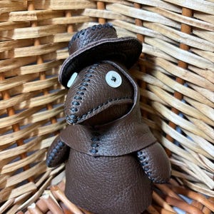 May include: A brown leather doll wearing a brown leather hat and a long brown leather coat. The doll has white button eyes and a stitched black mouth.