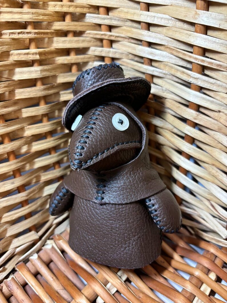 May include: A brown leather doll wearing a brown leather hat and a long brown leather coat. The doll has white button eyes and a stitched black mouth.