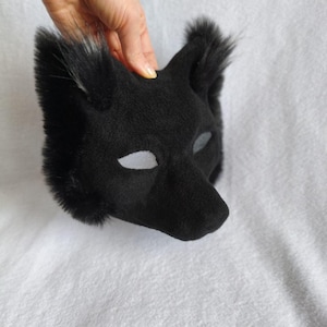 May include: A black plush animal mask with pointed ears and a snout. The mask has mesh eye openings and is held by a hand against a white background. The mask's fur has a soft texture.
