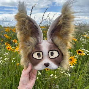 Può includere: Una maschera fatta a mano e stravagante che assomiglia alla faccia di un animale dei cartoni animati. La maschera presenta grandi occhi espressivi, un naso marrone e orecchie soffici e strutturate con un mix di pelliccia marrone e beige. La maschera è tenuta contro uno sfondo di fiori selvatici.