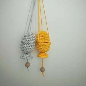 May include: Two crocheted egg-shaped pouches hanging from cords with wooden beads at the bottom. One pouch is silver and the other is yellow.