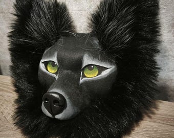 May include: A black wolf mask featuring long, dense black faux fur around the perimeter. The face is sculpted with a smooth matte finish, highlighting a black nose and striking, almond-shaped eyes with bright yellow-green irises and white accents.