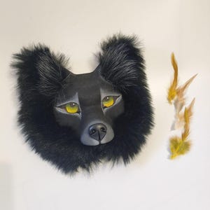 Peut inclure: Un masque de loup noir avec un visage sculpt&eacute; lisse, des yeux jaune vif et un nez noir, entour&eacute; d'une &eacute;paisse fourrure synth&eacute;tique noire. Trois petites plumes brun clair et jaunes sont plac&eacute;es sur le c&ocirc;t&eacute; droit du masque.