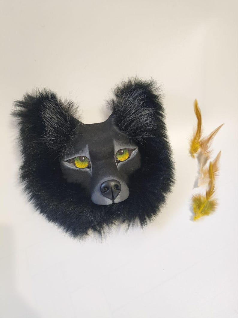 Peut inclure: Un masque de loup noir avec un visage sculpt&eacute; lisse, des yeux jaune vif et un nez noir, entour&eacute; d'une &eacute;paisse fourrure synth&eacute;tique noire. Trois petites plumes brun clair et jaunes sont plac&eacute;es sur le c&ocirc;t&eacute; droit du masque.