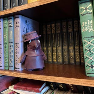 Può includere: Una statuina artigianale di medico della peste in pelle bordeaux scuro con cappello, in piedi su una libreria di legno. Lo scaffale è pieno di vari libri, alcuni con scritte dorate e copertine verdi.