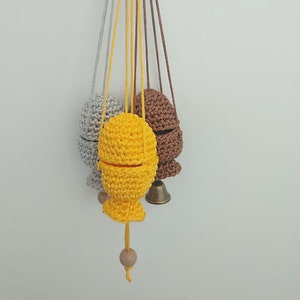 May include: Several handmade crochet fish-shaped ornaments in shades of yellow, grey, and brown. Each ornament is suspended by a string. Some have a wooden bead and a small bell attached. The ornaments are displayed against a white backdrop.