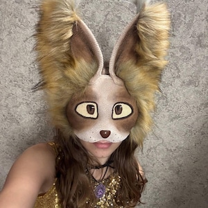 May include: A fox mask in shades of brown and tan, featuring large, fluffy ears with white tips. The mask has a detailed face with large eyes and a small nose. The mask is worn over the face.