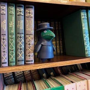 May include: A handcrafted leather plague doctor figurine with a green beak and a dark blue coat and hat. The figurine is displayed on a wooden shelf among a collection of vintage books with colorful spines.