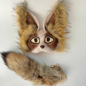 May include: A fox mask and tail set, ideal for costumes. The mask has large eyes, a brown nose, and long, fluffy ears. The tail is made of brown and tan faux fur, with a gold-coloured clasp. The set is designed for costume or cosplay.