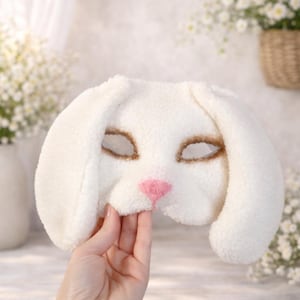 Könnte beinhalten: Eine weiße Plüsch-Hasenmaske mit großen, schlaffen Ohren. Die Maske hat braune Augenöffnungen und eine rosa Nase. Die Maske wird von einer Hand gehalten und zeigt ihre weiche Textur und ihr verspieltes Design.