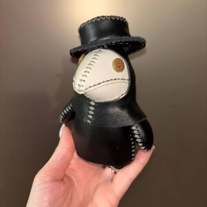 May include: A black leather plague doctor doll with a white beak and a black top hat. The doll has a red button eye.