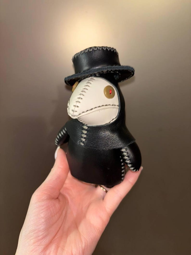 May include: A black leather plague doctor doll with a white beak and a black top hat. The doll has a red button eye.