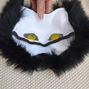Pu&ograve; includere: Una maschera da gatto bianca con occhi giallo brillante e pupille nere, circondata da un bordo spesso di lunga pelliccia sintetica nera. La maschera include un cinturino elastico nero fissato sopra l'area degli occhi per tenerla ferma.