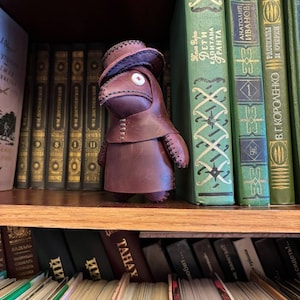 Könnte beinhalten: Handgefertigte, dunkelbraune Leder-Pestdoktor-Figur mit Hut und Mantel, die auf einem Holzregal vor einer Sammlung alter Bücher steht. Die Figur hat ein genähtes Design und ein weißes Kreuz auf ihrer Maske.
