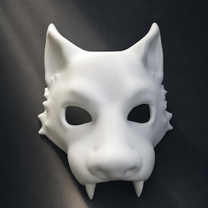 May include: A matte white wolf mask featuring pointed ears, almond-shaped eye openings, a prominent snout, and two sharp fangs extending downward from the upper jaw. The mask has a smooth, sculpted surface with textured fur details along the sides.
