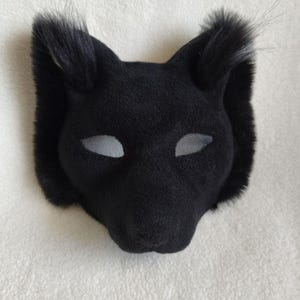May include: A black animal mask featuring soft, plush-like fabric covering the entire surface. The mask has pointed ears with tufts of longer black fur at the tips and almond-shaped eye openings covered with a fine, light grey mesh material.