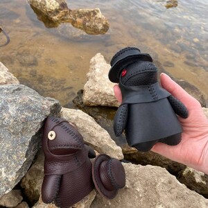 May include: Two small leather figures wearing hats. The figure on the left is brown and the figure on the right is black. Both figures have stitched details and button eyes.