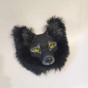 Peut inclure: Un masque de loup noir dot&eacute; d'un museau sculpt&eacute;, d'oreilles pointues et d'yeux jaune vif. Le masque est entour&eacute; d'une &eacute;paisse bordure en fausse fourrure sombre qui encadre le visage, avec de subtils reflets gris sur le bout des oreilles.