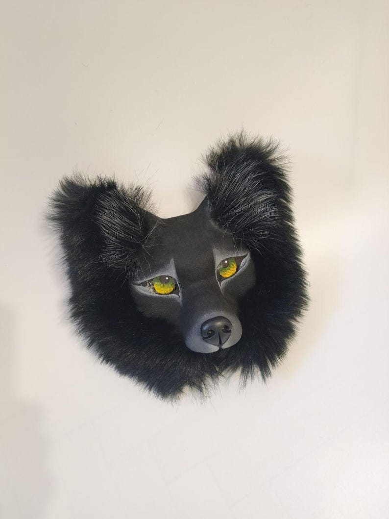 Peut inclure: Un masque de loup noir dot&eacute; d'un museau sculpt&eacute;, d'oreilles pointues et d'yeux jaune vif. Le masque est entour&eacute; d'une &eacute;paisse bordure en fausse fourrure sombre qui encadre le visage, avec de subtils reflets gris sur le bout des oreilles.