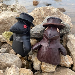 May include: Two small, handmade leather dolls wearing black and brown plague doctor masks with hats. The dolls are sitting on a pile of rocks near a body of water.