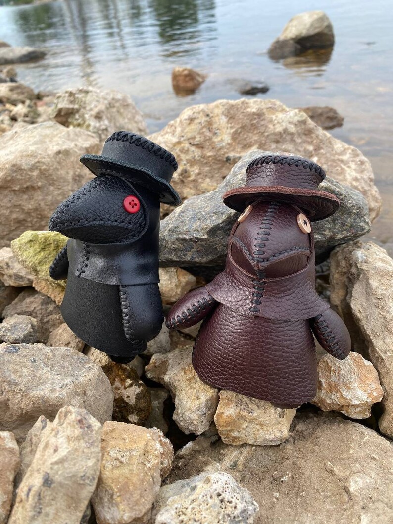 May include: Two small, handmade leather dolls wearing black and brown plague doctor masks with hats. The dolls are sitting on a pile of rocks near a body of water.
