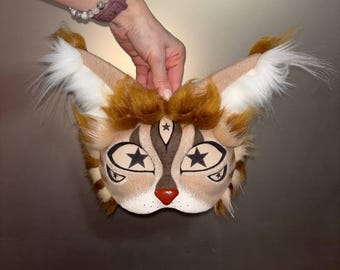 May include: A brown and white furry cat mask with black star-shaped eyes and a brown nose. The mask has a white fur trim around the edges.