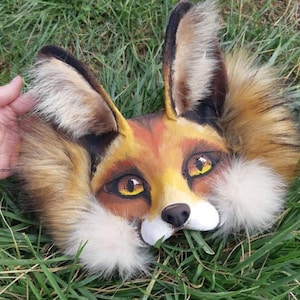 May include: A detailed fox mask with a hand-painted design. The mask has a brown, orange, and yellow face, large eyes, and soft faux fur around the ears and cheeks. The mask is resting on green grass.