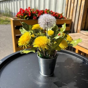 May include: A small, galvanized metal bucket filled with artificial dandelions. The arrangement includes bright yellow flowers, green leaves, and a fluffy, white dandelion seed head. The bucket sits on a black table, with red flowers in the background.