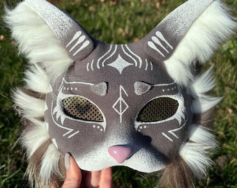 May include: A gray and white cat mask with white fur trim. The mask has a white design on the face and a pink nose. The eyes are covered with a mesh material.