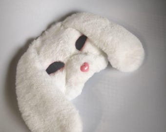 May include: A white plush rabbit mask featuring soft faux fur, large black oval-shaped eye openings, and a small, glossy pink nose. The mask has long, floppy ears extending from the sides and a rounded face shape with a textured, fuzzy surface.
