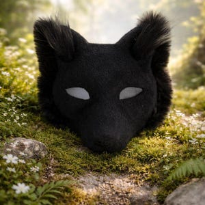 Puede incluir: Una máscara de zorro negro con cortes blancos para los ojos, sobre un fondo natural de musgo, rocas y follaje. La máscara tiene un aspecto texturizado y peludo, con orejas puntiagudas y un hocico definido. La estética general recuerda a una criatura del bosque.