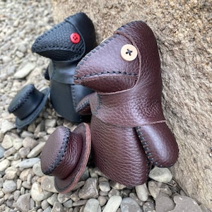 May include: Two handmade leather plague doctor dolls. One is black with a red button nose and the other is brown with a beige button nose. Both dolls are wearing hats.