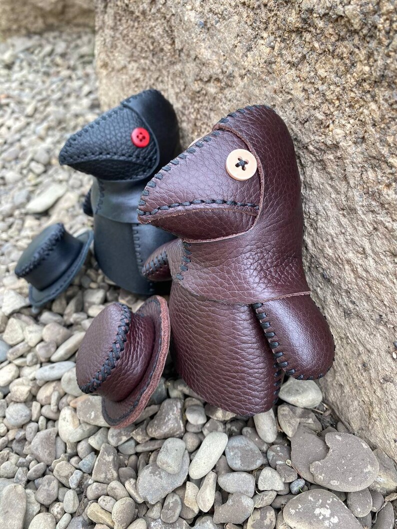 May include: Two handmade leather plague doctor dolls. One is black with a red button nose and the other is brown with a beige button nose. Both dolls are wearing hats.