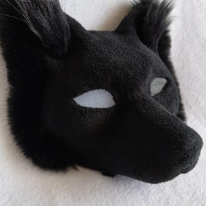 Op de afbeelding: Een zwart dierenmasker gemaakt van zachte, pluche stof met puntige oren en een snuit. Het masker is voorzien van witte gaasbedekkingen voor de ogen die zicht mogelijk maken. De textuur van het materiaal is pluizig, vergelijkbaar met vacht, en de algehele vorm is ontworpen om de kop van een hondachtig of wolfachtig wezen na te bootsen.