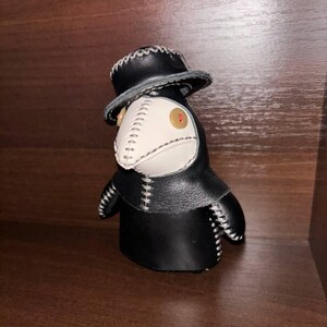 May include: A black leather plague doctor doll with a white beak and a black hat. The doll has white stitching around the edges of the beak, hat, and body.