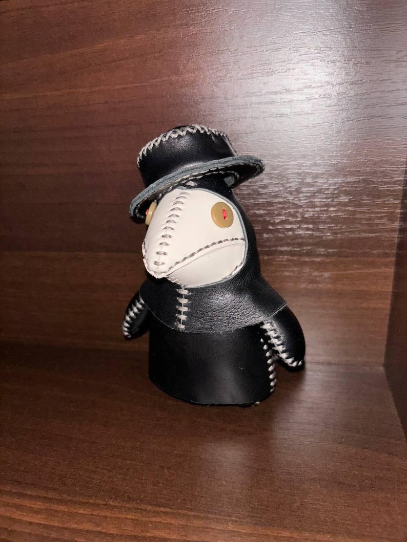 May include: A black leather plague doctor doll with a white beak and a black hat. The doll has white stitching around the edges of the beak, hat, and body.