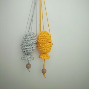 May include: Two hanging crochet egg-shaped ornaments. One is silver, the other is yellow. Each has a wooden bead at the bottom and a string for hanging. The yellow egg is open, revealing the inside. The ornaments are handmade.