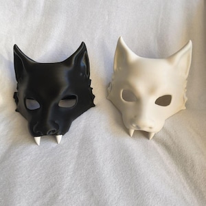 May include: Two wolf masks, one black and one off-white, are displayed side-by-side. Each mask features pointed ears, eye openings, and two small, white fangs. The masks are resting on a white, textured surface.