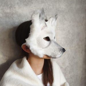 Op de afbeelding: Een wit wolvenmasker met textuurdetails die op vacht lijken, puntige oren met grijze uiteinden en een zwarte neus. Het masker bedekt het bovenste deel van het gezicht en wordt vastgezet met een band, met een realistisch dierlijk ontwerp in wit en grijs.