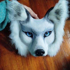 Pu&ograve; includere: Una maschera da lupo con pelliccia sintetica bianca e grigia, occhi blu brillante con eyeliner nero e puntini bianchi sopra gli occhi. La maschera presenta un muso scolpito con un naso nero e orecchie a punta con le estremit&agrave; grigie.