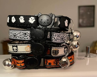 May include: A set of four Halloween-themed cat collars with black and white patterns, including ghosts, spiders, pumpkins, and skulls. Each collar has a safety breakaway buckle and a bell.