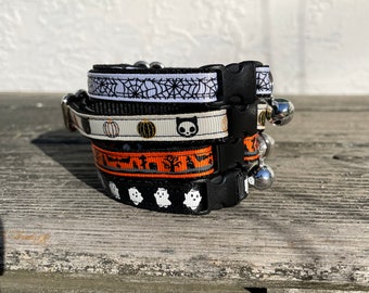 Pode incluir: Um conjunto de quatro coleiras de gato pretas com diferentes padrões temáticos de Halloween, incluindo abóboras, fantasmas e teias de aranha. Cada coleira tem um sino de prata.