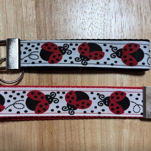 May include: Two keychains with a white background and black and red ladybugs. The top keychain has a black ribbon and the bottom keychain has a red ribbon.