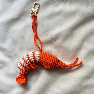 May include: A handmade crochet shrimp keychain. The shrimp is orange with a white and orange striped tail. It has a gold-colored clasp and orange string for attachment. The keychain is a whimsical accessory.